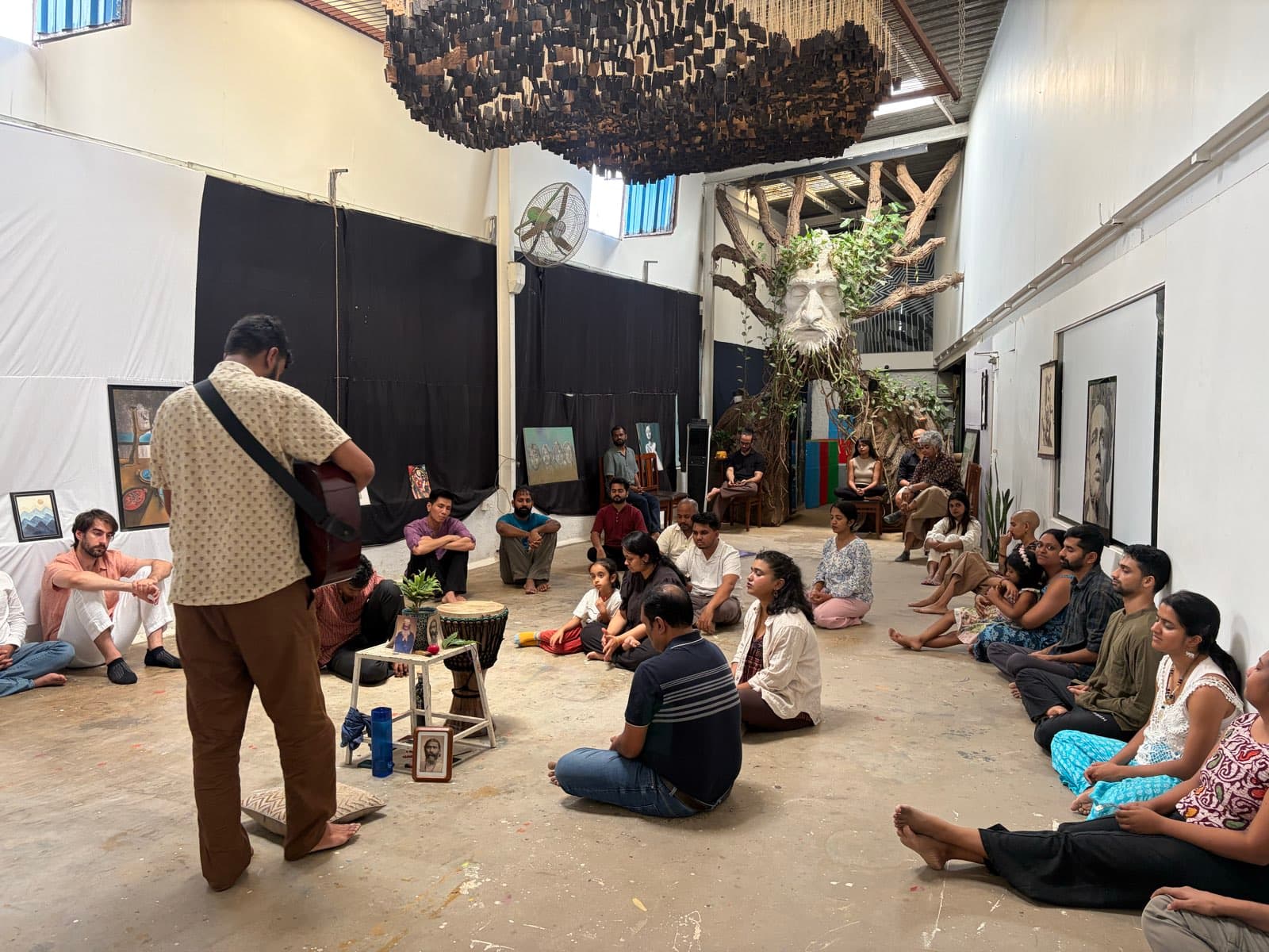 People sitting in a loose circle on the warehouse floor while a musician plays guitar among them.