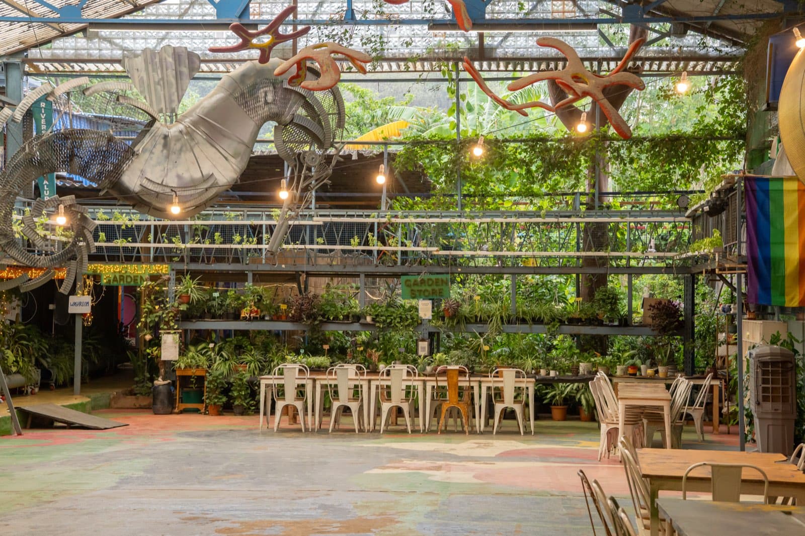 The open warehouse floor under a glass roof, ringed with plants, a large sculptural bird hanging overhead.